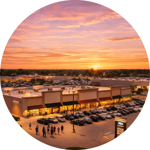 Sunset view over a busy shopping plaza with lit storefronts and a crowded parking lot.