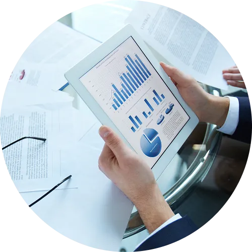 Person holding a tablet displaying blue financial bar and pie charts, with documents and glasses on a glass table.